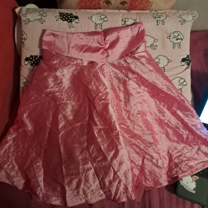 Rosa långkjol med rosett och volang (barn) - Supersöt rosa långkjol i glansigt tyg med bred midja och stor rosett bak. Kjolen har volangdetaljer och flera lager för extra fluffig känsla. Perfekt för dig som vill sticka ut med en feminin och lekfull stil. (Botten gör att den kan hålla sin form)