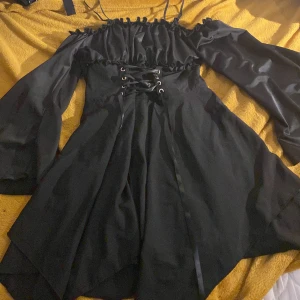 Svart offshoulder klänning med snörning - Supersnygg svart kortklänning med offshoulder och volangdetaljer upptill. Klänningen har långa vida ärmar, smala axelband och snörning i midjan för extra edge. Kjolen är lätt utsvängd och har lager på lager-effekt. Perfekt för dig som gillar en unik och trendig look. (Ny med lapp kvar) 