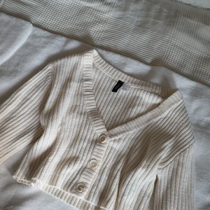 Beige ribbad kofta från Divided - Säljer en croppad, ribbstickad kofta från Divided i beige. Koftan har v-ringning, tre stora knappar framtill och långa ärmar. Perfekt att slänga över ett linne eller t-shirt för en chill och snygg look.