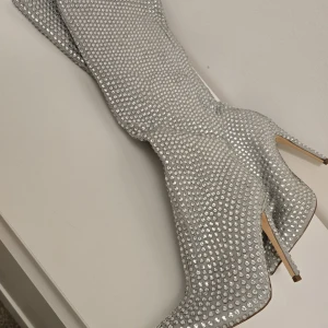 Glittriga silverstövletter med klack - Säljer ett par riktigt snygga stövletter i silver med glittriga stenar över hela skon. De har spetsig tå och hög klack, perfekta för att sticka ut. Materialet är syntet och de är täckta av glittriga detaljer som ger en lyxig känsla.