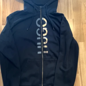 Svart hoodie från HUGO med dragkedja - Snygg svart hoodie från HUGO med silverfärgat logotryck framtill. Tröjan har huva med snörning, hel dragkedja och praktisk känguruficka. Perfekt för en avslappnad och cool stil.