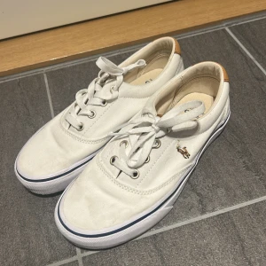 Vita sneakers från Polo Ralph Lauren - Fräscha vita sneakers från Polo Ralph Lauren med klassisk design. Skorna har snörning, bruna detaljer i skinn på hälen och Polo-logga bak. Ovandel i bomullstyg och vit sula med blå rand. Perfekta för en clean och stilren look. Endast använda 4 gånger.