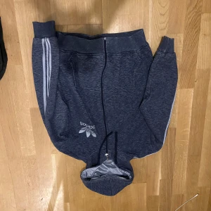 Mörkgrå Adidas mjukisbyxor L - Mörkgrå mjukisbyxor från Adidas med klassiska vita ränder längs sidorna och logga på låret. Byxorna har resår och dragsko i midjan samt muddar vid bensluten. Perfekta för chill eller träning, riktigt sköna och stilrena.