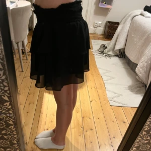 Svart volangkjol - Säljer en svart kortkjol med volanger och bred resår i midjan. Kjolen har ett flowigt och lätt tyg som ger en snygg siluett. Perfekt för dig som gillar en feminin och trendig stil.