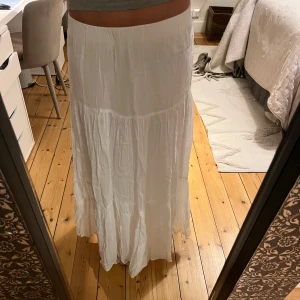 Vit långkjol i bomull - Luftig och flowy vit långkjol med lätt rynkade partier och bred resår i midjan. Kjolen har en bohemisk känsla och är perfekt för varma dagar. Tillverkad i tunt, mjukt bomullstyg som ger en avslappnad och skön vibe.