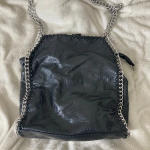 Svart axelväska med kedjedetaljer Tiamo - Cool svart axelväska från Tiamo i skinnimitation med grova silverfärgade kedjor längs kanterna. Väskan har dragkedja upptill och en extra ficka med dragkedja på baksidan. Perfekt för dig som gillar edgy detaljer och vill ha plats för allt du behöver.