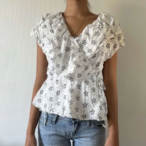 Vit omlotts topp med blommigt mönster - Söt vit omlotts topp med svart blommigt mönster och volangdetaljer vid axlarna. Toppen har korta ärmar, knyt i sidan och lätt rynkad midja!🌸