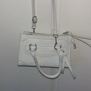 Vit axelväska i skinnimitation - Snygg vit axelväska i skinnimitation med justerbar axelrem och två handtag. Väskan har en dragkedja upptill och en mindre ficka med dragkedja på framsidan. Stilren och enkel design med silvriga metalldetaljer. Perfekt storlek för det viktigaste.
