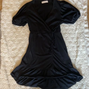Svart omlottklänning Samsa Collection - Supersnygg svart omlottklänning från Samsa Collection med puffiga korta ärmar, knyt i midjan och volangkant nertill.  Klänningen har v-ringning och är i mjukt material💗