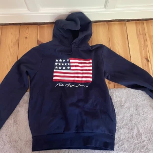 Marinblå hoodie Polo Ralph Lauren  - Snygg marinblå hoodie från Polo Ralph Lauren i bomull. Stor amerikansk flagga framtill och vit broderad logga under. Klassisk huva och långa ärmar, perfekt för en chill och sportig look.