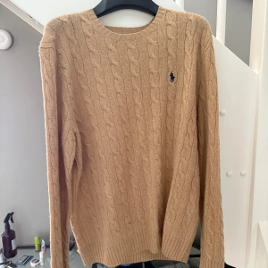 Beige kabelstickad tröja Ralph Lauren - Kabelstickad beige tröja från Ralph Lauren med klassisk rund halsringning och broderad marinblå logga på bröstet. Tröjan har lång ärm och ribbade muddar, perfekt för lager-på-lager under kyliga dagar.