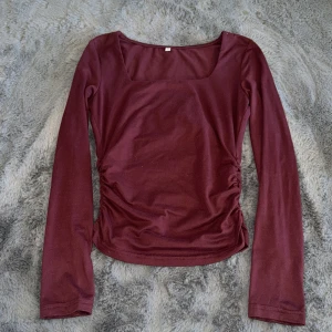 Vinröd långärmad topp  - Snygg vinröd långärmad topp med fyrkantig urringning och rynkade sidor. Toppens material är mjukt och stretchigt, perfekt för en trendig look. Passformen är slim och den har en enkel men stilren design.