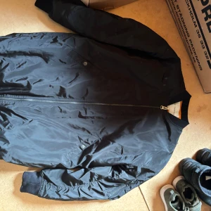 Svart bomberjacka från NLY Trend - Svart bomberjacka från NLY Trend i storlek 34. Jackan har klassisk bomberstil med ribbade muddar, dragkedja framtill och orange foder. Snygg och enkel att matcha till olika outfits, perfekt för dig som gillar streetstyle. Jackan är lång i kroppen vilket ger en unik look! 