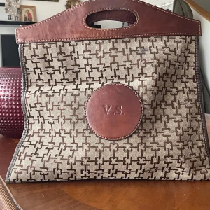 Beige och brun shoppingväska V.S. - Stilren shoppingväska från Victoria’s Secret i beige och brunt med grafiskt mönster och detaljer i brunt skinn. Väskan har utskurna handtag och en rund skinnpatch med V.S. på framsidan. Perfekt för dig som gillar en unik och klassisk look.