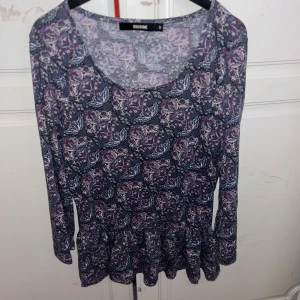 Blommig långärmad topp från Bik Bok - Supersöt långärmad topp från Bik Bok i storlek S. Mörkblå botten med blommigt mönster i lila, rosa och vitt. Rund halsringning och volang nertill ger en extra feminin touch. Perfekt att styla med jeans eller kjol för en trendig look.