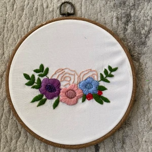 Broderad Tavla  - Bilden visar en broderad tavla med färgglada blommor i en rund träram. Broderad hemma.