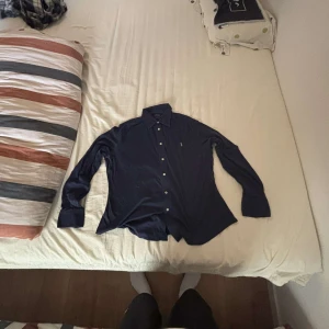 Mörkblå skjorta från Polo Ralph Lauren - Snygg mörkblå skjorta från Polo Ralph Lauren i klassisk modell med knappar framtill och lång ärm. Skjortan har en stilren look och är tillverkad i mjukt material som känns skönt mot huden. Perfekt för dig som gillar en clean och tidlös stil.