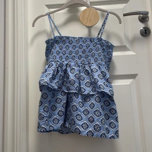 Blå mönstrad kortklänning Gina Tricot - Supersöt blå och vit topp från Gina Tricot med smala axelband och volangdetalj. Toppen har ett snyggt mönster och smock upptill för en bekväm passform. Perfekt för varma dagar och somriga vibes!❤️storlek 170