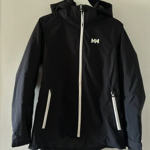 Täckjacka - En svart täckjacka/skidjacka med vita detaljer från Helly Hansen i storlek M. I fint skick. Skriv för mer bilder. 