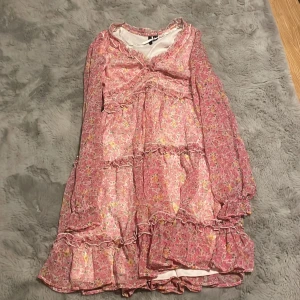 Blommig rosa kortklänning Vero Moda - Supersöt kortklänning från Vero Moda i rosa med blommigt mönster och volangdetaljer. Lätt och flowy modell med långa ärmar och v-ringning. Perfekt för vår och sommar när du vill känna dig extra fin.