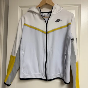 Nike Tech Hoodie - Nike Tech hoodiejacka i vitt med grå och gula detaljer samt svart logga på bröstet. Jackan har hel dragkedja, huva och två fickor. Ärmarna har coola färgblock i gult och grått. Perfekt för dig som gillar sportig och avslappnad stil. Har några fläckar på ärmarna men jag har inte provat att få bort dom med tex Galltvål eller Vanish som nog skulle fungerar om man vill pröva att få bort dom