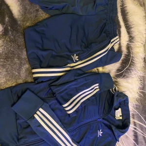 Blå Adidas träningsset XS - Säljer ett klassiskt blått Adidas träningsset i storlek XS. Setet består av en zip-jacka och matchande byxor med vita ränder längs ärmar och ben samt Adidas logga på bröst och lår. Materialet är mjukt och följsamt, perfekt för sport eller chill. En vacker tracksuit för endast 200kr