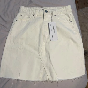 Vit jeanskjol från Gina Tricot - Vit jeanskjol från Gina Tricot med rå kant nertill, klassiska femficksdetaljer och bälteshällor. Kjolen har en rak passform och är kort, perfekt till sneakers eller boots. Snygg och enkel att styla med crop top eller hoodie.