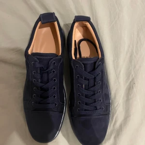 Mörkblå sneakers från Christian Louboutin - Snygga mörkblå sneakers från Christian Louboutin i mocka med klassisk röd sula och broderad logga på hälen. Skorna har rund tå, platt sula och snörning. Perfekta för dig som vill sticka ut med exklusiva detaljer och lyxig känsla. Skolåda finns att få med.