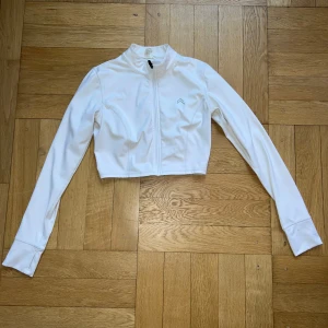 Vit croppad zip-tröja Oneractive. - Snygg vit croppad tröja med lång ärm och dragkedja framtill från Oneractive. Tröjan har en sportig look med hög krage och är tillverkad i ett stretchigt, slätt material som känns bekvämt mot huden. Perfekt för dig som gillar en clean och modern stil. Org 650kr
