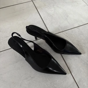 Svarta klackar med spetsig tå från H&M - Stilrena svarta pumps från H&M med smal klack och spetsig tå. Skorna har öppen häl med smal rem runt ankeln och är tillverkade i blankt syntetmaterial. Perfekta för dig som gillar en klassisk och elegant look.