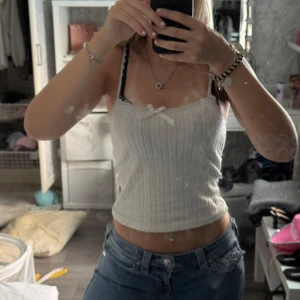 Vit ribbad linnetopp med spets - Supersöt vit ribbad linnetopp med tunna axelband och spetskant. Toppen har en liten rosett framtill och ett diskret mönster i tyget som ger en extra touch. Perfekt att styla med jeans eller kjol för en fräsch look.