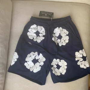 Blå shorts med blommigt tryck Denim Tears - Mörkblå shorts från Denim Tears med stora vita blommiga tryck över hela plagget. Elastisk midja med snörning och en bakficka. Perfekta för sommaren och riktigt snygga till en enkel t-shirt eller hoodie.