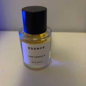 ESSNCE I Am Vanilla Eau de Parfum - Parfym med söt och krämig vaniljdoft, toppad med orkidé och jordgubbe. Hjärtnot av tonkaböna och basnoter av mysk, brunt socker och amberwood. Kommer i en rund glasflaska med svart lock och stilren vit etikett.