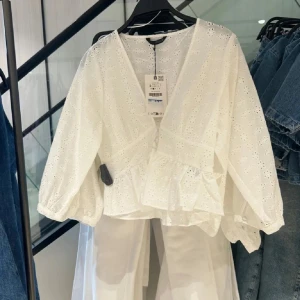 Vit blus med broderade blommor - Superfin vit blus med v-ringning och broderade blommor. Blusen har trekvartsärmar med resår vid ärmslut, omlottkänsla och volang nertill. Perfekt till jeans för en fräsch och somrig look.