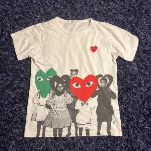 Comme des Garçons Play t-shirt  - TVÄRFET‼️ Vit t-shirt från Comme des Garçons Play med ikoniska hjärtmotiv i rött, grönt och svart framtill. Trycket föreställer barn med hjärtformade masker och ett litet rött hjärta på bröstet. Klassisk passform och rund hals. 100% bomull. Hör av er vid minsta fundering🙌