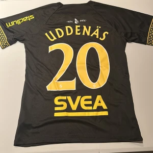 AIK matchtröja Uddenäs #20 Nike - AIK:s matchtröja med Uddenäs och nummer 20 på ryggen. Billig då stadium-printet är upp och nedvänt. Svart bas med gula detaljer, klubbmärke på bröstet och sponsorer som Truecaller och Svea. Korta ärmar med unika mönster vid ärmslut. Tillverkad i lätt och ventilerande funktionsmaterial. Replika. Endast handtvätt gäller!