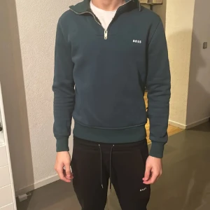 Mörkgrön half zip tröja från BOSS - Snygg mörkgrön tröja från BOSS med half zip och hög krage. Tröjan har lång ärm, ribbade muddar och diskret BOSS-logga på bröstet. Perfekt för dig som gillar stilren och sportig look.
