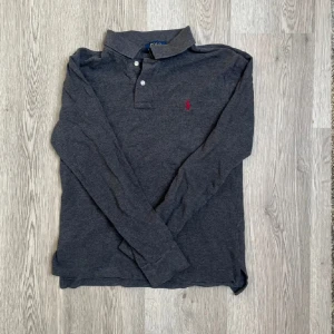 Mörkgrå pikétröja från Polo Ralph Lauren - Snygg mörkgrå långärmad pikétröja från Polo Ralph Lauren med klassisk krage och knäppning framtill. Tröjan motsvarar en S-Xs, kom gärna med frågor🤩🤝