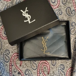Svart YSL korthållare - Snygg svart korthållare från YSL med ikoniskt guldigt logospänne framtill. Korthållaren är tillverkad i strukturerat skinn med quiltad design och har en elegant, stilren look. Perfekt för dig som vill ha en lyxig accessoar med statement-känsla.