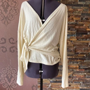 Ribbad omlottopp från NLY Trend - Krämvit ribbad omlottopp från NLY Trend i storlek S. Toppen har långa ärmar, v-ringning och knyt i sidan. Knapp i ena sidan. Sömmen har släppt lite på ett ställe, lätt att laga (se bild)