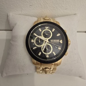 Versus Versace guldig klocka med svart urtavla - Snygg klocka från Versus Versace med guldig länk och svart urtavla. Klockan har flera små urtavlor, datumvisning och detaljer i guld. Länken pryds av ett lejonhuvud och spännet har Versus Versace-loggan. Perfekt accessoar för dig som gillar statement.