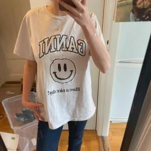Ljusrosa GANNI t-shirt med smiley - Vit t-shirt från GANNI med svart tryck framtill, stor smiley och texten 'Have a nice day!'. Klassisk rund halsringning och avslappnad passform. Perfekt för dig som gillar coola statement-plagg och enkel stil.