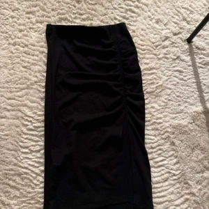 Svart långkjol med slits och rynk - Säljer en svart långkjol med snygg rynkdetalj längs ena sidan och en slits framtill som ger kjolen en cool vibe. Kjolen är figurnära och stretchig, perfekt att styla med sneakers eller boots. Passar dig som gillar minimalistisk och stilren look. Jättebra skick.
