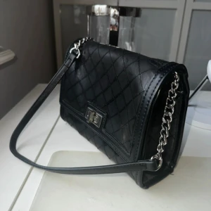 Svart quiltad axelväska med kedja - Snygg svart axelväska i quiltad design med silverfärgad kedja och vridlås. Väskan har en stilren look med rutmönster och lång axelrem i både skinn och metall. Perfekt storlek för det viktigaste och ger en classy vibe till din outfit.