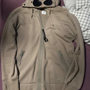Beige hoodiejacka från C.P. Company - Säljer en beige hoodiejacka från C.P. Company med ikoniska goggles i huvan. Jackan har dragkedja framtill, två fickor med dragkedja och ribbade muddar. Perfekt för dig som vill sticka ut med streetwear-vibe.