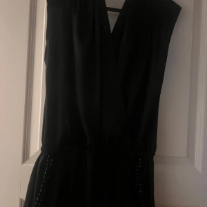 Svart jumpsuit från Lipsy - Supersnygg svart jumpsuit från Lipsy med knyt bakom nacken och markerad midja. Den  har coola detaljer med svarta nitar vid fickorna och är tillverkad i ett glansigt, mjukt material som känns lyxigt.