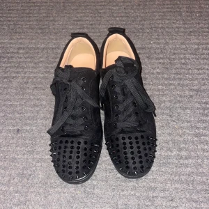 Svarta sneakers med nitar från Louboutin - Pris kan ändras vid snabbt köp! Svarta sneakers i mocka från Christian Louboutin med coola svarta nitar på tån och klassisk röd sula. Skorna har snörning och platt sula, vilket ger en edgy vibe. Perfekta för dig som vill sticka ut med en lyxig streetstyle.