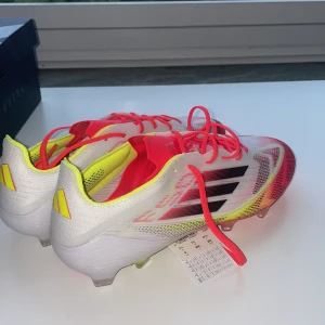 Adidas F50 Elite FG fotbollsskor - Säljer ett par Adidas F50 Elite FG fotbollsskor i storlek 42. Skorna har en snygg design i vitt, neonrosa och gult med svarta detaljer och snörning. Ovandelen är i syntetmaterial och sulan har fasta dobbar för gräsplan. Perfekta för dig som vill sticka ut på planen! Diskuterar gärna pris!