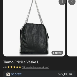 Svart axelväska Tiamo Priscilla - Säljer en svart axelväska från Tiamo med  silverfärgade kedjor som axelrem och detaljer längs kanterna. Väskan är gjord i syntetmaterial med en grynig läderlook, har dragkedja både på utsidan och insidan samt flera fack för småprylar.