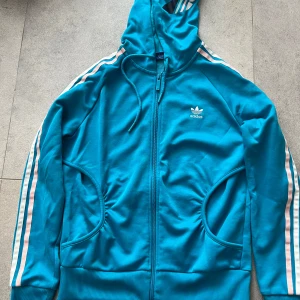 Blå Adidas vindjacka med huva - Säljer en blå Adidas vindjacka med huva och dragkedja. Klassiska vita och rosa ränder längs ärmarna och stort Adidas-tryck på huvan. Jackan har två fickor och ribbade muddar. Perfekt för dig som gillar sportig och retro stil.